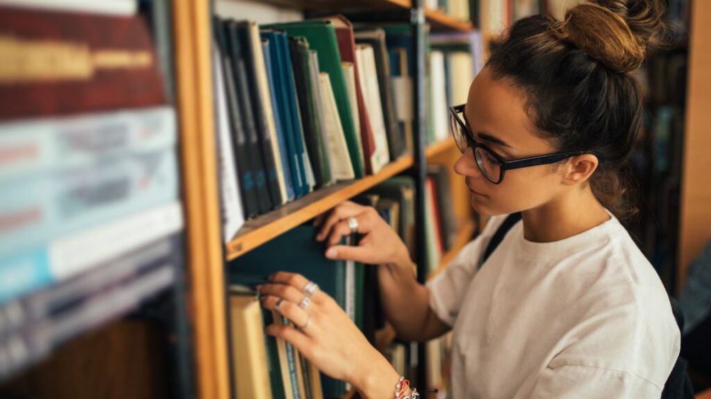 Uma jovem de óculos e cabelos presos em um coque examina atentamente os livros em uma estante de biblioteca ou livraria, tocando a lombada de um exemplar.