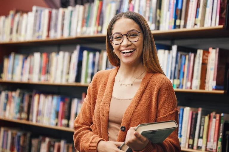 Mulher negra sorri para foto segurando um livro em uma biblioteca.
