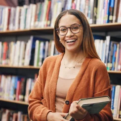 Mulher negra sorri para foto segurando um livro em uma biblioteca.